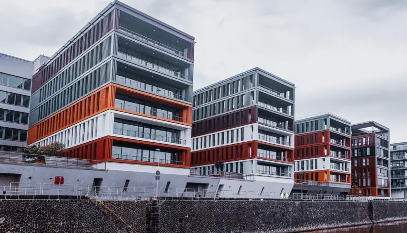 Nowoczesne budynki o geometrycznych kształtach, z elementami w kolorze pomarańczowym i bordowym, tworzą futurystyczny krajobraz miasta nad wodą.
