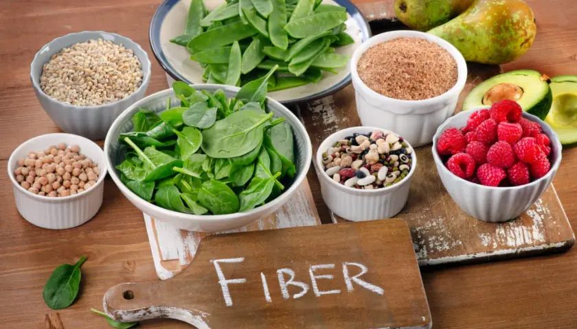 En skärbräda med texten "FIBER" omgiven av skålar med bönor, linser, spenat, hallon, päron, avokado och korn.