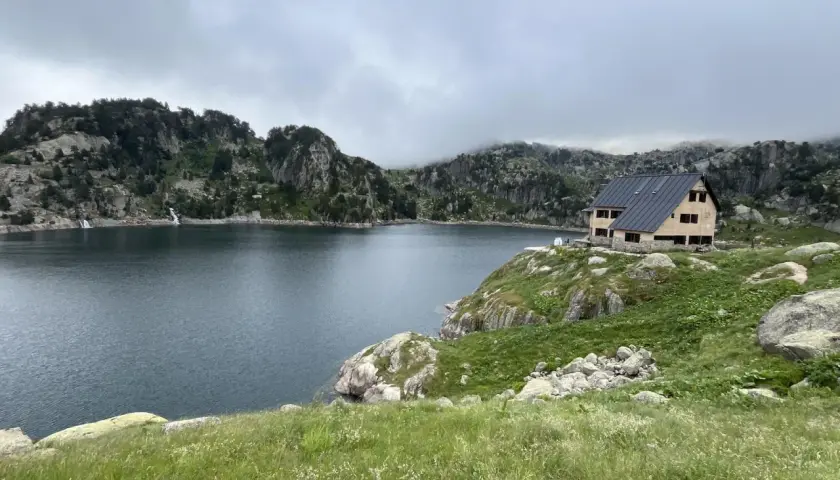 Refugio de montaña junto a un lago rodeado de montañas rocosas y vegetación.