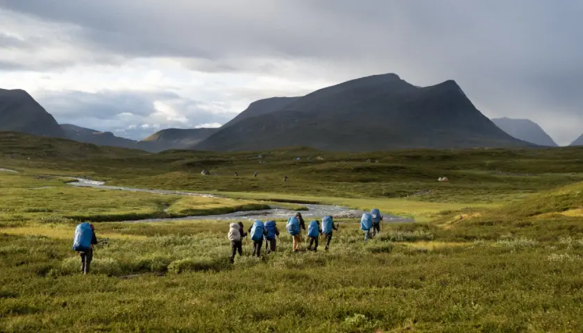 Hikers with blue backpacks trek through a vast, green landscape in Sweden, reminiscent of the Fjällräven Classic. Tents dot the distant hills under a dramatic sky.