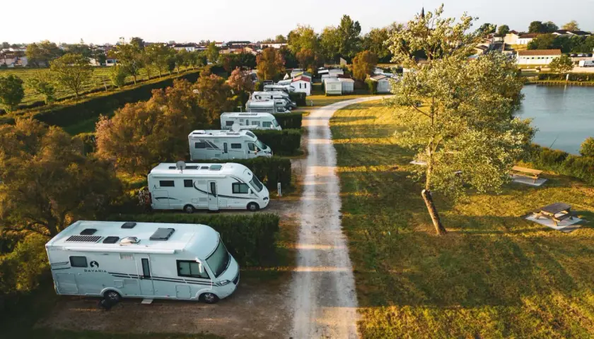 Vue aérienne d'un air camping car park, avec plusieurs camping-cars blancs alignés le long d'un chemin de terre, près d'un lac et d'une petite ville.