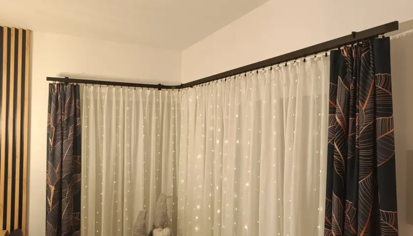 Elegancki karnisz apartamentowy czarny, na którym wiszą zasłony z motywem liści i delikatne firanki ze światełkami.