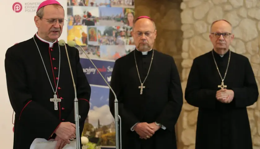 Trzech biskupów w czarnych szatach i z krzyżami na szyjach podczas zjazdu biskupów. Jeden z nich przemawia przy mikrofonie.