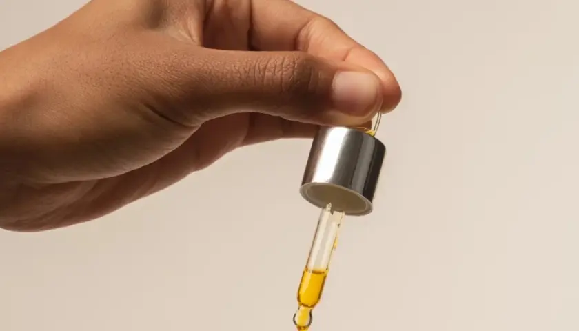 Dłoń trzyma pipetkę z kroplą złotego oleju buriti, gotową do aplikacji.
