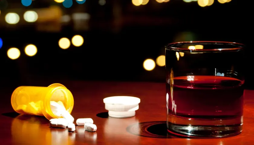 Nalany ibuprofen i szklanka alkoholu na drewnianym blacie, w tle rozmyte światła.