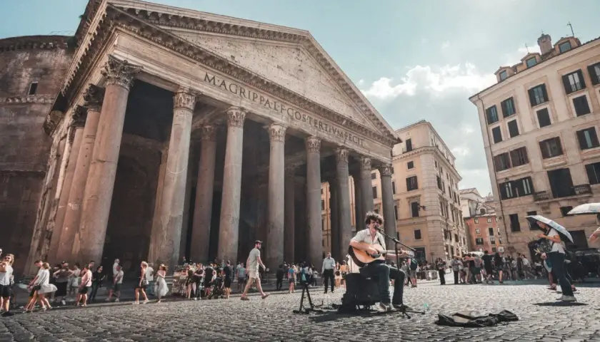 Un musicien joue de la guitare devant le Panthéon d'Agrippa, un monument historique de Rome, tandis que des touristes se promènent sur la place pavée.