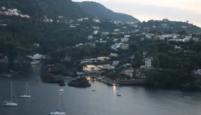 Vittoria Colonna a Ischia: case aggrappate alla collina, luci soffuse e barche a vela nella baia al crepuscolo.