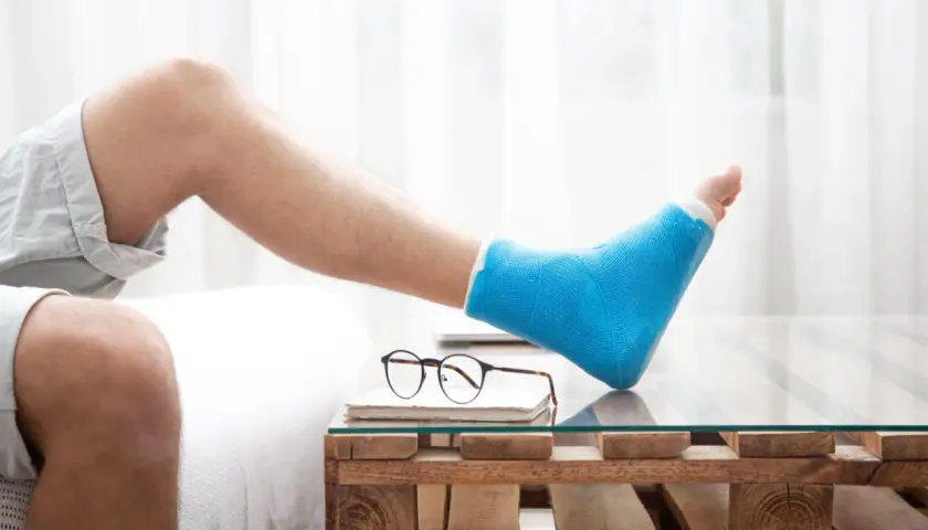 Mann mit blauem Gipsbein ruht sich aus. Die Frage "welche schuhe nach fersenbeinbruch" beschäftigt ihn, während er auf einem Holztisch mit Glasplatte entspannt.