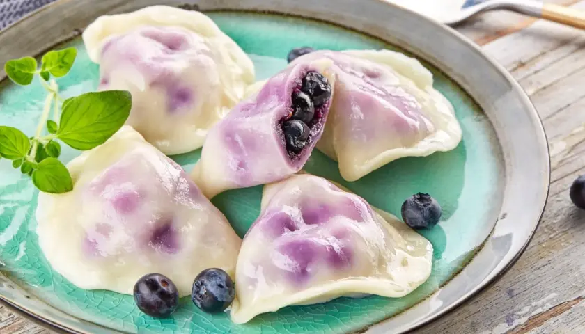 Pyszne pierogi z jagodami, jeden przekrojony, ukazujący soczyste owoce. Podane na turkusowym talerzu z gałązką ziół.