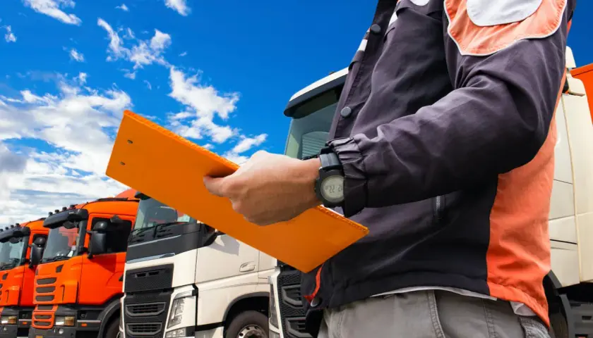 Kierowca z teczką, na której widnieje świadectwo kwalifikacji zawodowej, stoi przed rzędem ciężarówek.