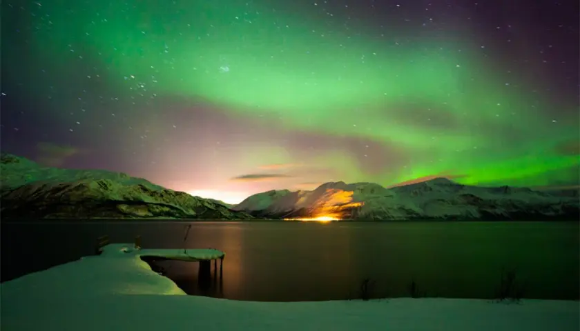 Spektakuläre Nordlichter über schneebedeckten Bergen und einem gefrorenen See. Ein perfekter Winterurlaub in Skandinavien.