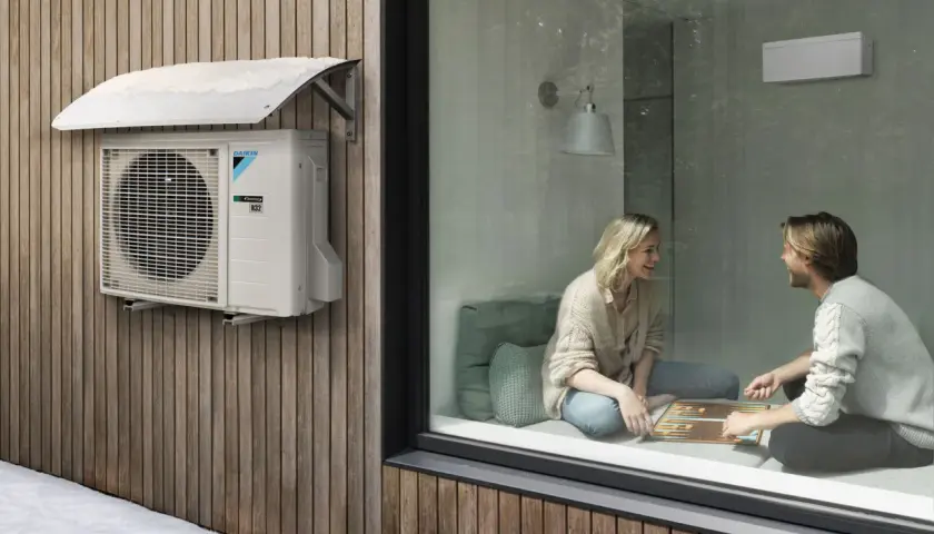 Unidad exterior de aire acondicionado Daikin instalada con la distancia adecuada a la pared de madera. Dentro, una pareja juega.