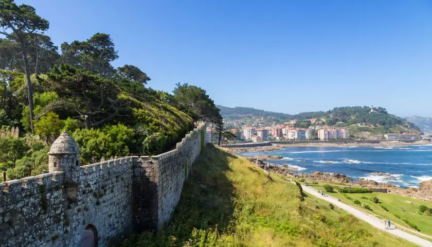 A historic stone wall winds along the coast of Baiona, Portugal, overlooking the blue sea and a town nestled on the hills.