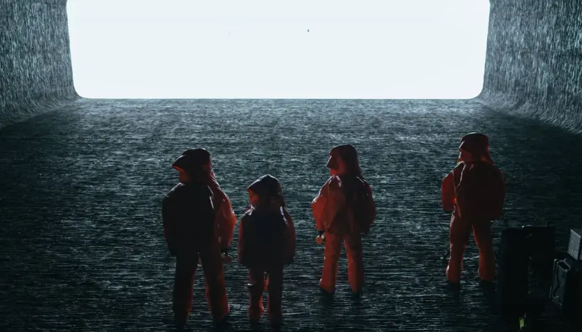 Persone in tute arancioni osservano un'enorme apertura luminosa, evocando scene di "Arrival" e la sua spiegazione di un evento alieno.
