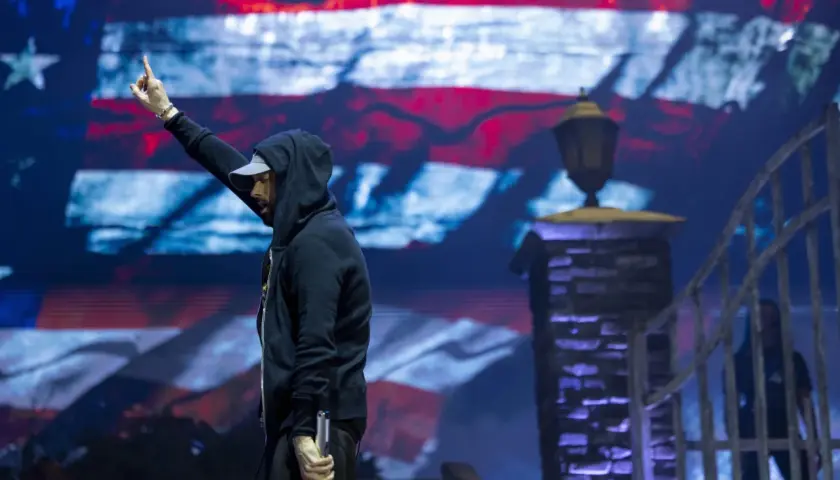 Eminem na koncercie w Polsce, z ręką uniesioną w geście pozdrowienia, na tle ekranu z amerykańską flagą.