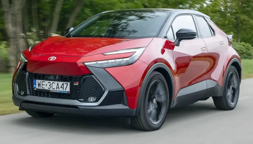 Czerwona Toyota C-HR 2020 z czarnym dachem pędzi po drodze, otoczona zielenią drzew.