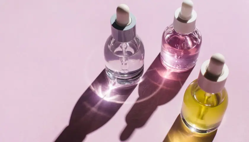 Trzy buteleczki z pipetką: przezroczysta, różowa i żółta. Cienie i refleksy światła na różowym tle. Czy zawierają parabeny w kosmetykach?
