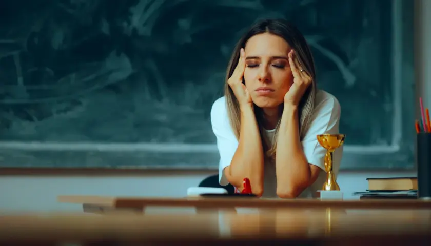Lehrerin mit Händen an den Schläfen, Augen geschlossen, wirkt erschöpft. Ein Pokal und Bücher auf dem Tisch deuten auf den Schulalltag hin, der zu Burnout bei Lehrern führen kann.
