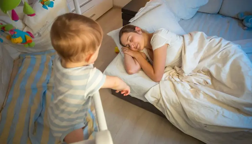 Un neonato di 7 mesi, in piedi nella sua culla, osserva la mamma addormentata nel letto.