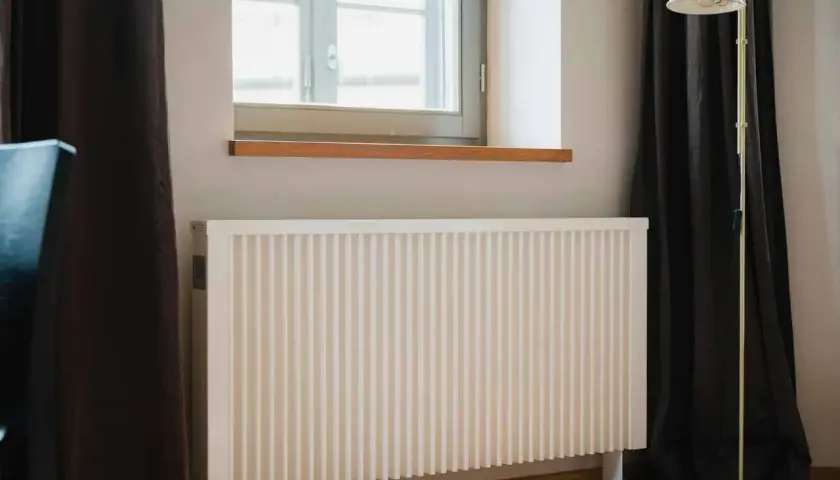 Biały kaloryfer pod oknem, obok ciemnej zasłony i lampy stojącej. Dowiedz się, jak ustawić kaloryfery przy ogrzewaniu gazowym dla optymalnego komfortu.
