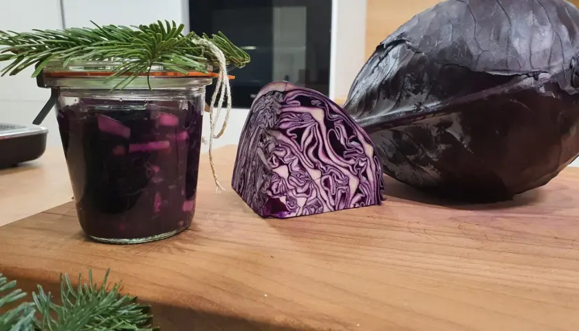 Ein Glas mit fermentiertem Rotkohl, daneben ein halber und ein ganzer Rotkohl. Tannenzweige schmücken das Glas.