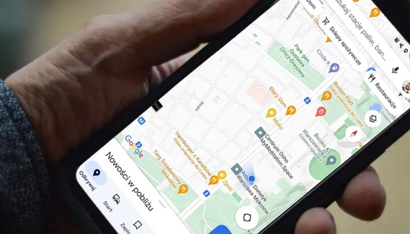 Dłoń trzyma telefon z mapą Google. Widoczna jest opcja udostępnianie lokalizacji, a także pobliskie restauracje i sklepy.