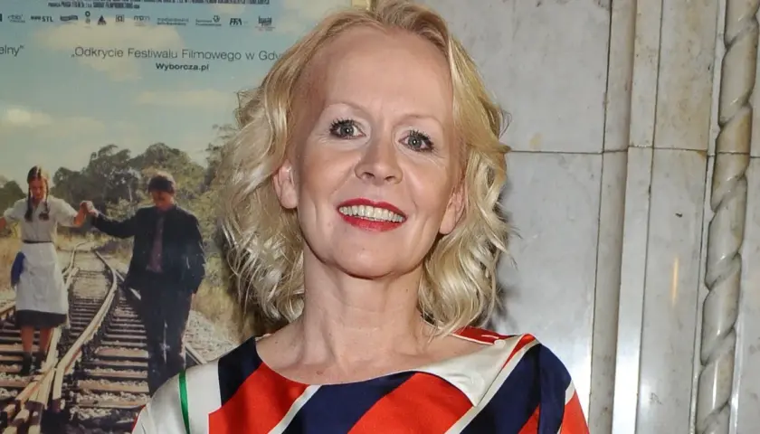 Agnieszka Krukówna uśmiecha się, ubrana w kolorowy strój. W tle plakat filmowy z parą na torach.