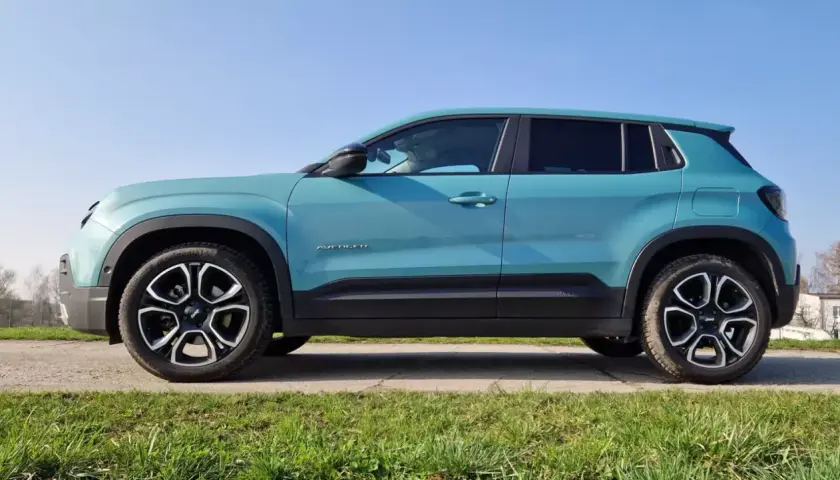 Turkusowy Jeep Avenger widziany z boku na tle nieba i zielonej trawy.