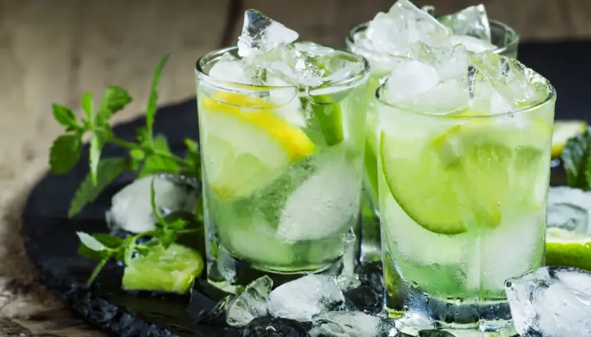 Trois verres de caipirinha glacée, garnie de citron vert et de citron, sur un plateau noir avec de la menthe fraîche. La recette parfaite pour se rafraîchir.