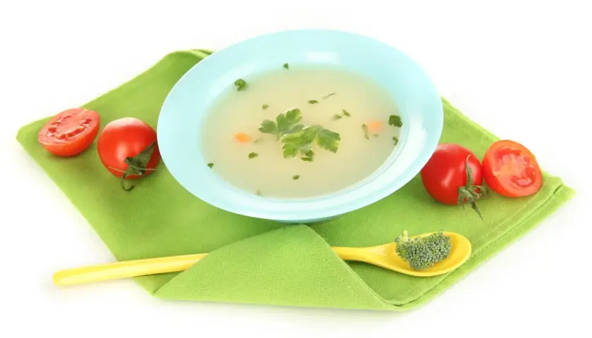 Brodo vegetale neonati in ciotola azzurra, con pomodorini e un cucchiaino giallo con broccoli.