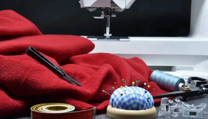 Czerwony materiał, maszyna do szycia, szpilki, miarka i nożyczki – wszystko gotowe do tworzenia patchwork projektów.