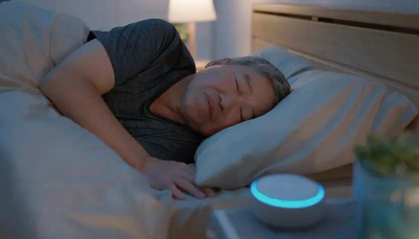 Ein Mann schläft tief und fest im Bett. Neben ihm leuchtet ein Smart Speaker, der ihm hilft, lange schlafen zu können.