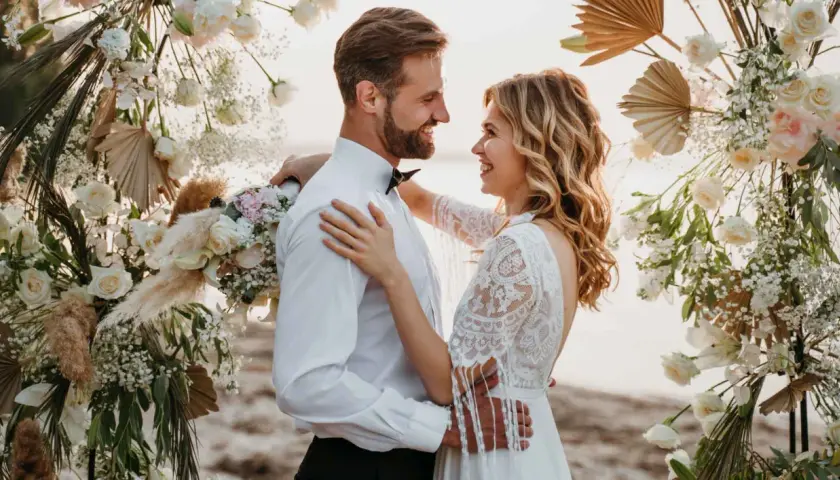 Un couple rayonnant s'embrasse sous une arche fleurie, célébrant leur mariage sans enfant sur une plage.