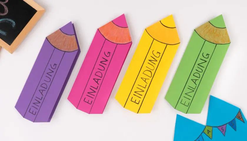 Bunte Stift-Bastelvorlagen mit "Einladung" beschriftet. Eine kleine Tafel mit ABC und eine blaue Wimpelkette sind ebenfalls zu sehen.