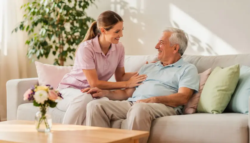 Eine Pflegerin spricht lächelnd mit einem älteren Mann auf einem Sofa. Ein Gespräch über den Eigenanteil im Pflegeheim kann so angenehm sein.