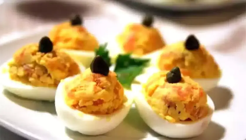 Six oeufs mimosa saumon garnis d'une olive noire sur une assiette blanche.
