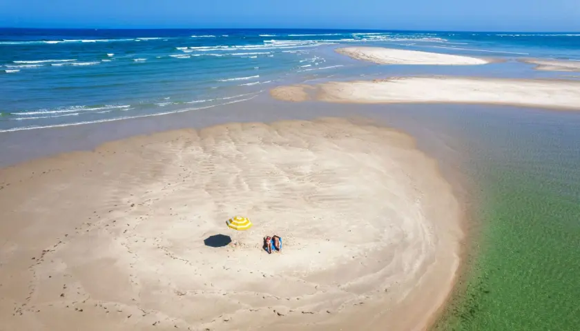 Dos personas descansan bajo una sombrilla a rayas en una playa de arena en Ria Formosa. El mar azul y las aguas cristalinas rodean la isla.
