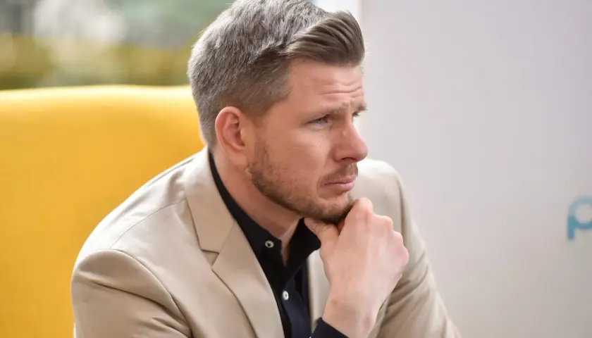 Mężczyzna w beżowej marynarce i granatowej koszuli, z ręką na brodzie, zastanawia się nad pytaniem: ile lat ma Filip Chajzer?