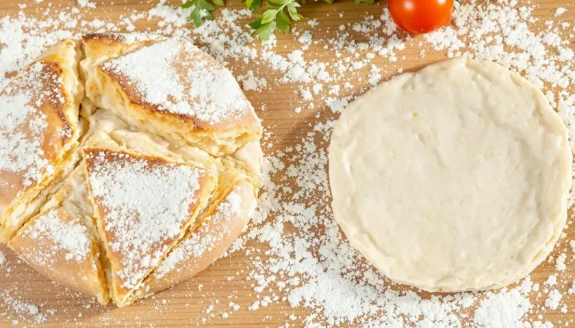 Une recette de pain maison : une miche dorée saupoudrée de farine à côté d'une pâte à pain crue, avec des tomates et du persil en arrière-plan.