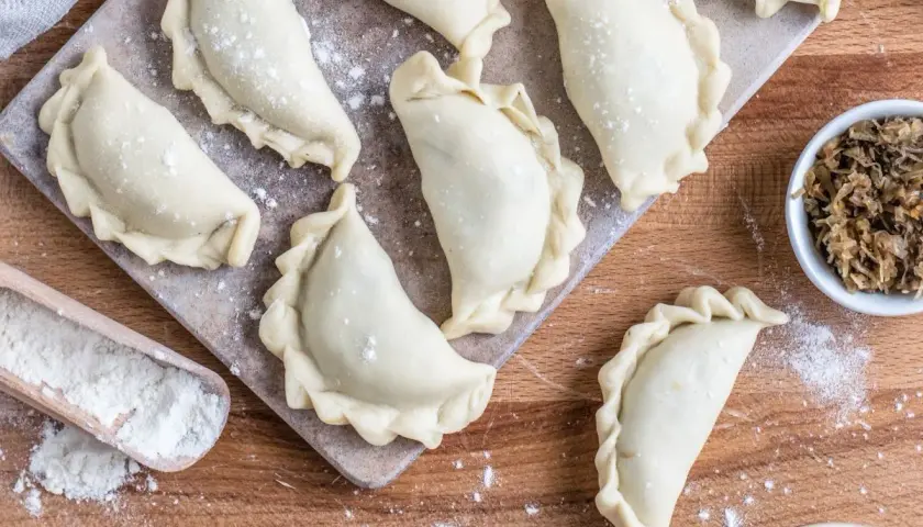 Surowe pierogi ruskie ułożone na desce, gotowe do zamrożenia. Obok miseczka z farszem i drewniana łopatka z mąką.