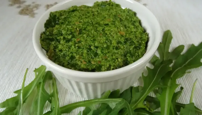 Pesto z rukoli w białej miseczce, otoczone świeżymi liśćmi rukoli. Idealne do makaronu lub jako dodatek do kanapek.
