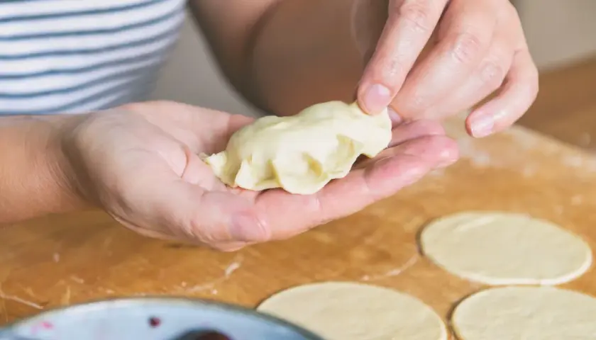 Dłonie formują pieroga z ciasta na pierogi bez jajka. Na blacie leżą okrągłe placki z ciasta.