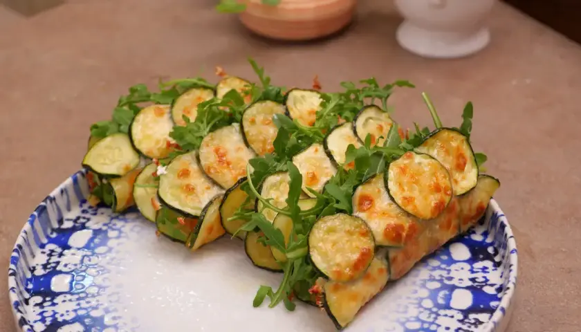 Rotolini di zucchine e rucola, cotti in padella e gratinati con formaggio fuso, serviti su un piatto decorato.