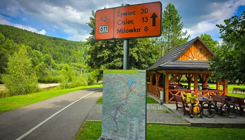Znak szlaku rowerowego Velo Sola z odległościami do Żywca, Ciśca i Milówki. Obok mapa i wiata.