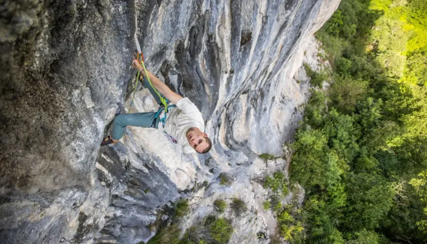 Un grimpeur escalade une paroi rocheuse, concentré sur sa prochaine prise. La végétation luxuriante contraste avec la pierre grise.