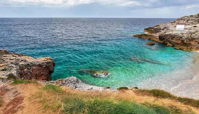 Vue d'une crique aux eaux turquoise cristallines, typique d'une plage de Pula en Sardaigne. Rochers et végétation méditerranéenne bordent la petite plage de galets.