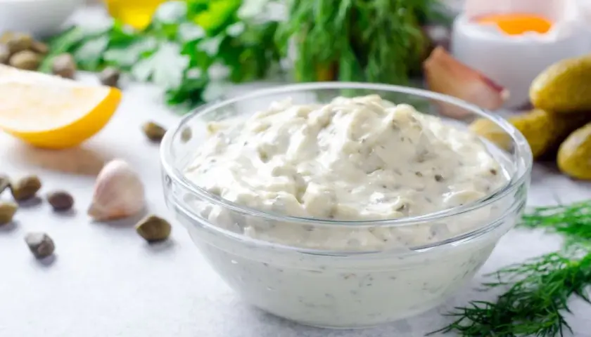 Una ciotola di cremosa salsa tartara, con capperi, limone, aglio e aneto sullo sfondo.
