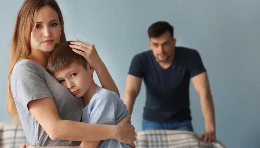Smutna kobieta tuli syna, podczas gdy w tle stoi zmartwiony mężczyzna. Trudno jej zaakceptować dziecko partnera.