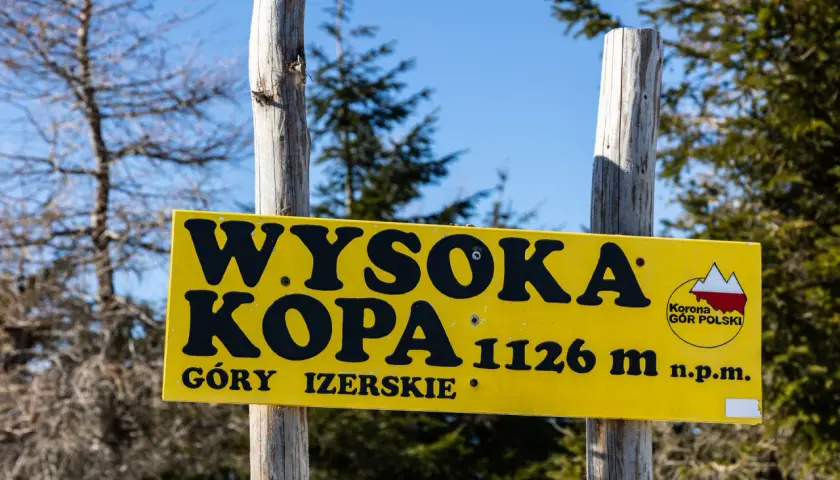 Żółta tablica na dwóch drewnianych słupkach informuje: "Wysoka Kopa 1126 m n.p.m. Góry Izerskie".