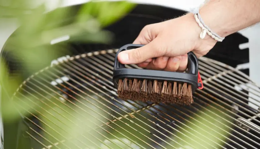 Main tenant une brosse pour nettoyer la grille du barbecue. Les herbes vertes floutées ajoutent une touche de fraîcheur à la scène.